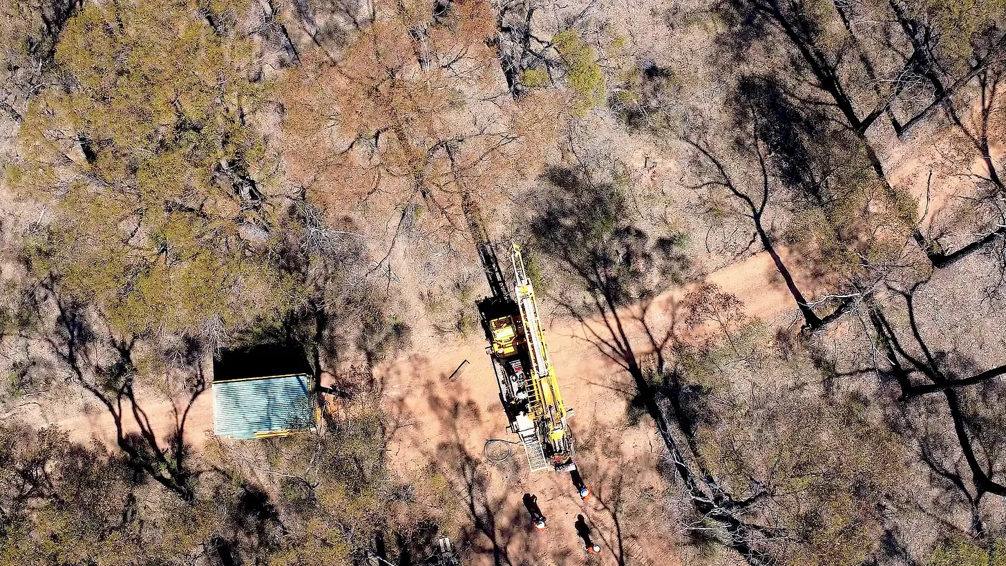 A yellow drill rig on a dirt track in dry bushland with workers in hard hats standing nearby.