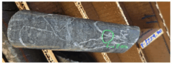 Image of Drill Core A close-up photograph of a cylindrical drill core sample resting in a cardboard core tray. The rock is dark grey and features a network of thin, white veins running through it. On the right side of the core, a specific area is circled in bright green marker, with handwritten green text underneath that appears to start with "Cu" (likely indicating copper). A wooden depth marker block with blue writing is partially visible in the background on the right.