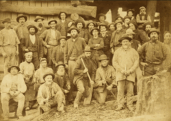 Vintage sepia photo of a large group of gold miners posing together with tools outside a timber building.