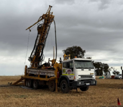 Drill rig mounted on a truck operating in a dry paddock under cloudy skies with trees and equipment in the background.