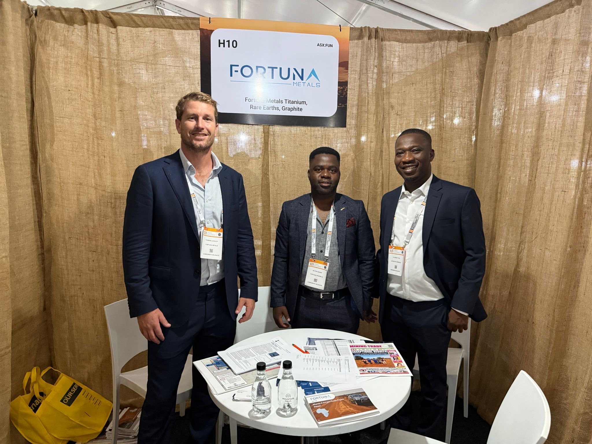 Three suited men at Fortuna Metals booth H10 with brochures and signage at mining conference.