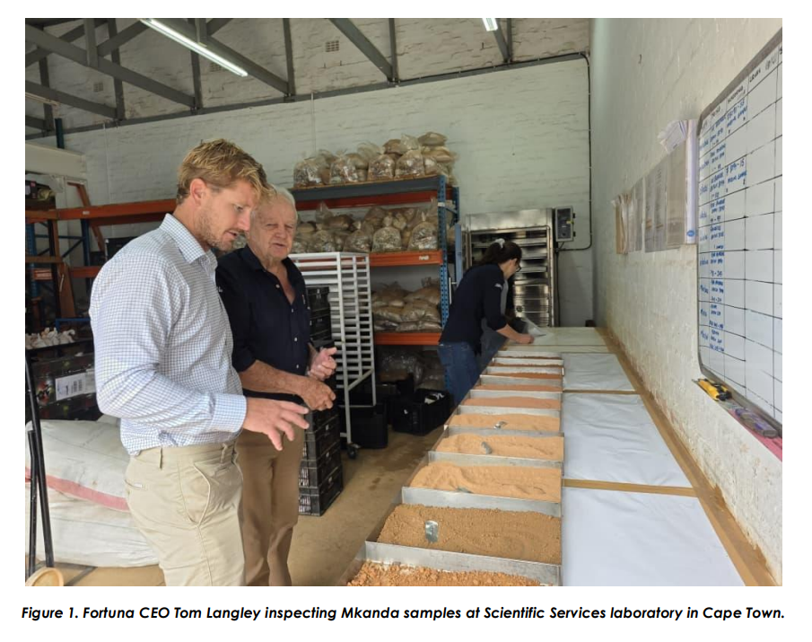 Two men inspect drill sample trays in a laboratory, with racks of bagged samples behind them.