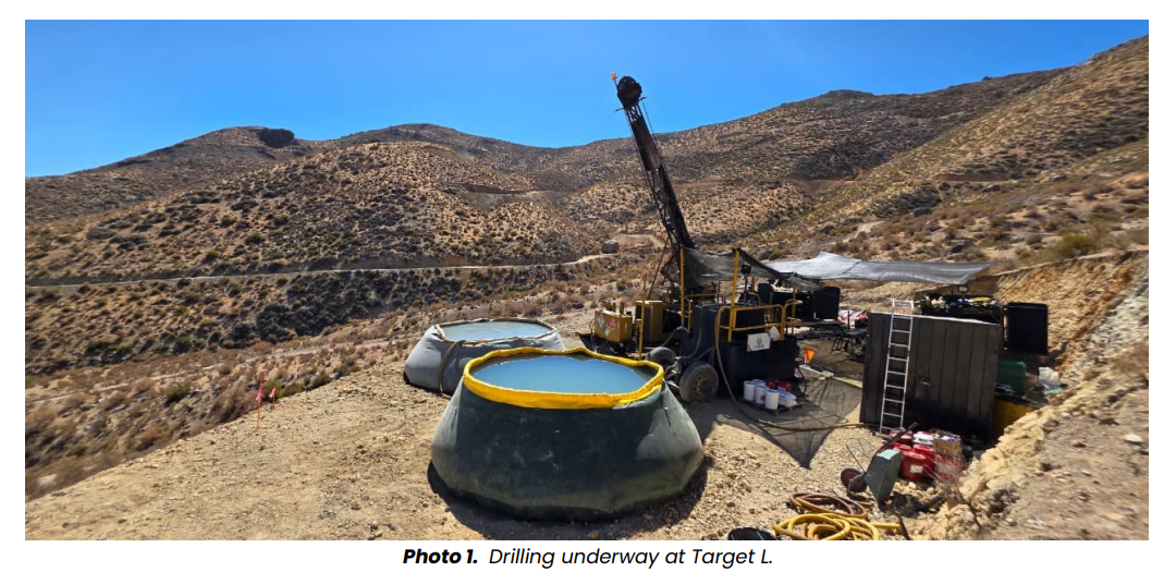 Drilling rig operating at Target L exploration site in arid hills, with water tanks, equipment and rugged terrain surrounding the drill pad.