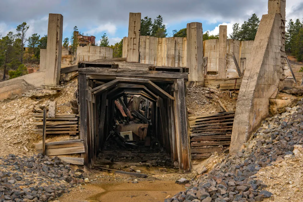 Abandoned mine entrance with timber supports, concrete ruins, rubble, and overgrown vegetation under cloudy sky
