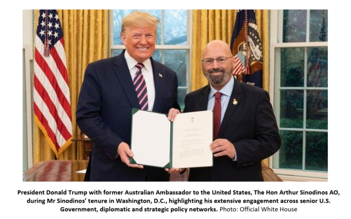 Former Australian Ambassador Arthur Sinodinos stands beside then-US President Donald Trump in the Oval Office, holding an official document.