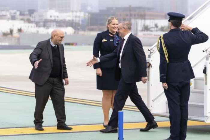 Arthur Sinodinos arrives on an airport tarmac, greeting officials beside an aircraft staircase while a uniformed officer salutes.