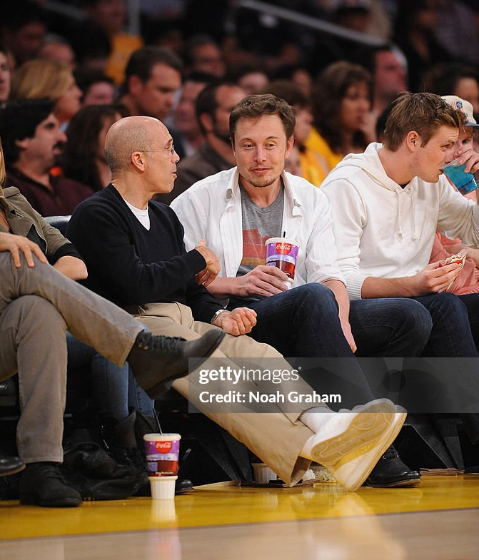 Jeffrey Katzenberg sits courtside at an NBA game, chatting with Elon Musk while spectators watch from the packed arena behind them.
