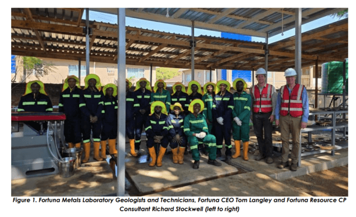 Group of Fortuna Metals geologists, technicians and executives at a laboratory site, wearing safety gear beneath a covered work area.