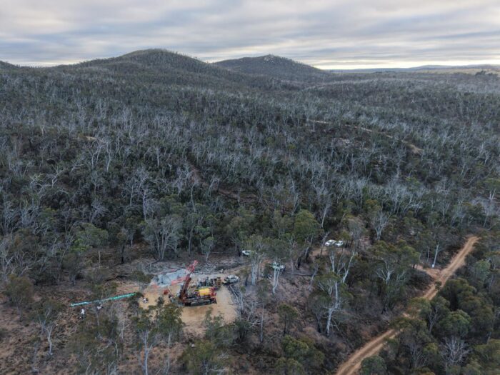 Aerial view of a drill rig operating at a cleared site within dense bushland, surrounded by rolling forested hills.