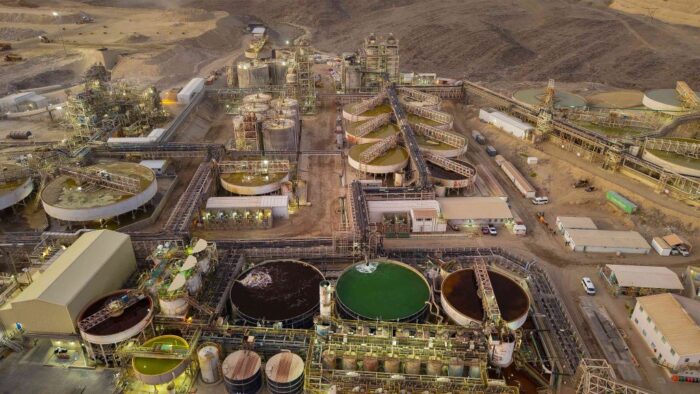 Aerial view of a large mineral processing plant with tanks, pipelines, conveyors and tailings facilities in a desert landscape.