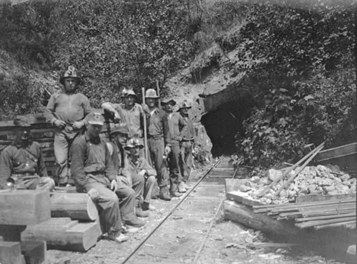 Historic black-and-white photo of miners gathered outside a mine entrance, with rail tracks leading into the tunnel and ore carts nearby.