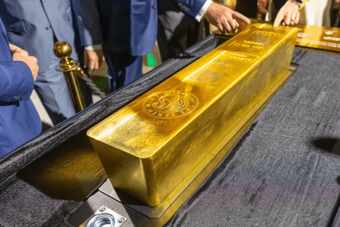 Large polished gold bar displayed on a table, with people gathered around it at an exhibition or event.