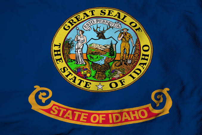 Flag of Idaho featuring the state seal on a blue background with a red and gold banner reading “State of Idaho”.