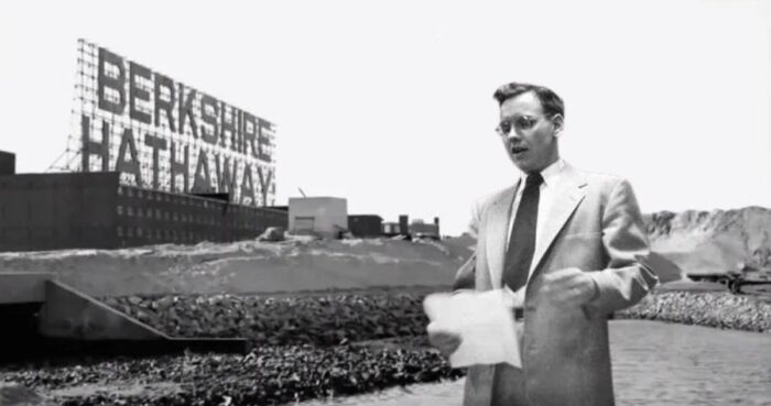 Black-and-white image of a man in a suit standing at an industrial site beneath a large Berkshire Hathaway sign.