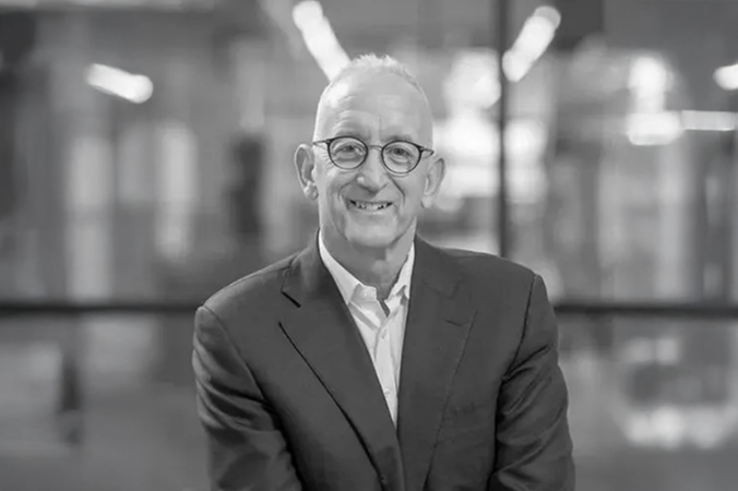 Black-and-white portrait of a smiling man wearing round glasses and a suit, seated in a modern office.