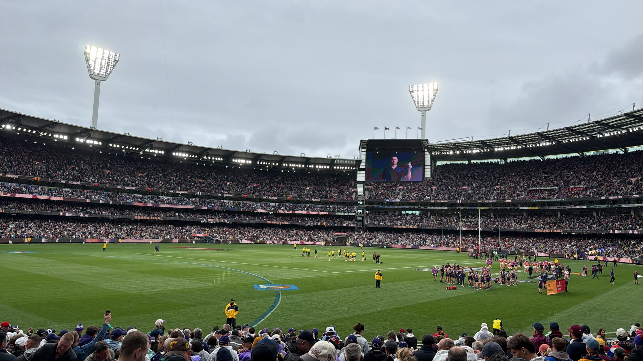 View from the stands at a packed MCG during the AFL Grand Final, with players gathered on the oval beneath floodlights and an overcast sky.