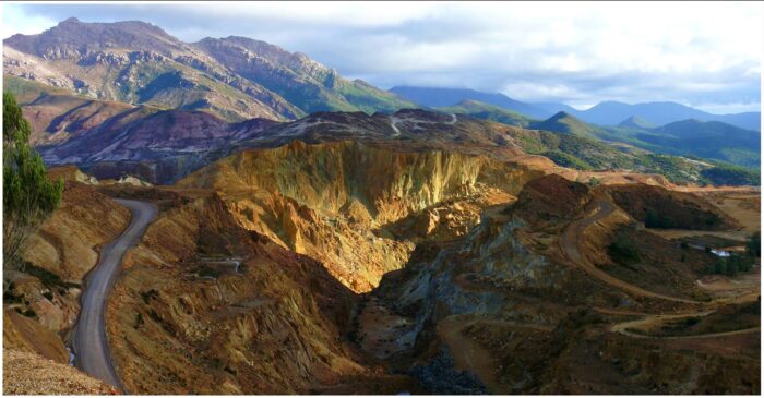 A vast open-cut mine with steep terraced rock faces, winding access roads and rugged mountains beneath a cloudy sky.