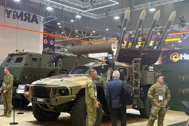 Military personnel stand beside armoured vehicles and a missile launcher system at an indoor defence industry exhibition.